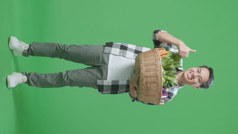cuerpo lleno de granjero masculino asiático que lleva una canasta de verduras mostrando los pulgares hacia arriba gesto y sonriendo mientras está de pie en el estudio de pantalla verde