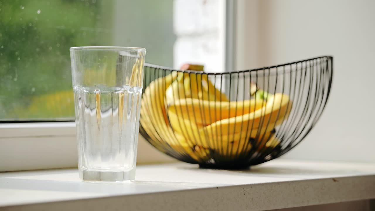 Three ice cubes drop into empty glass beside bananas in bright kitchen light