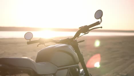 Close-up-footage-of-motorcycle-parking-on-the-ground-close-to-the-water-in-sunset,-sunlight-under-water-on-background