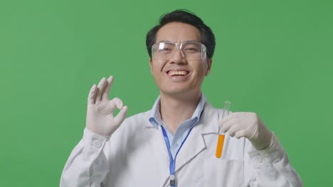 close up of asian man scientist with orange liquid in the test tube smiling and showing okay gesture while standing on the green screen background in the laboratory