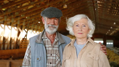 portrait of happy old caucasian married couple of farmers looking and smiling at camera while standing in stable with sheep flock