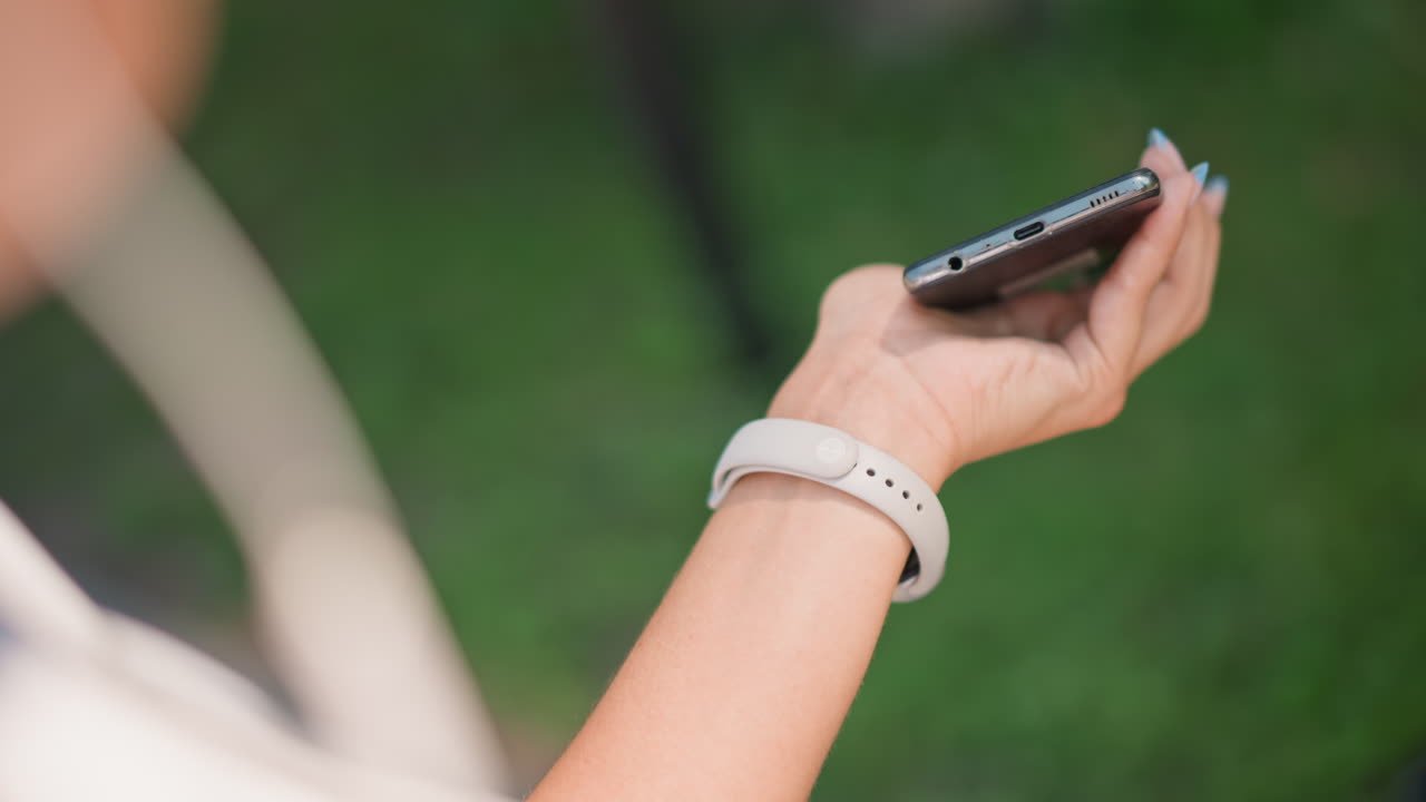 케이블이 연결된 스마트폰을 머리 위에서 손으로 잡고 있고, 콘텐츠 제작자가 재생 목록을 스크롤하고 있으며, 손목시계가 보이고, 햇빛이 나뭇잎 사이로 비치고, 자연스러운 매니큐어, 부드러운 보케 녹색 배경, 평화로운 분위기