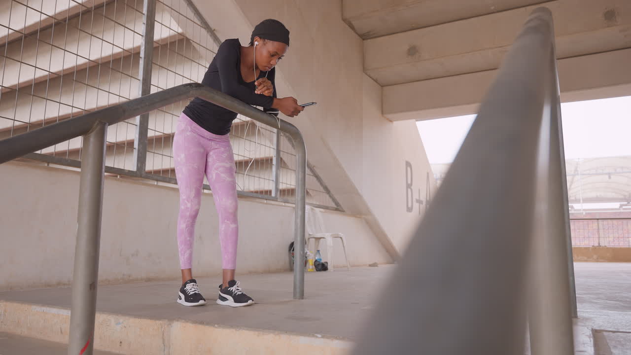 young woman checking phone on stairwell, leaning on metal railing under concrete overpass, wearing pink pants and sneakers while scrolling messages quiet urban scene with architectural stairs, soft