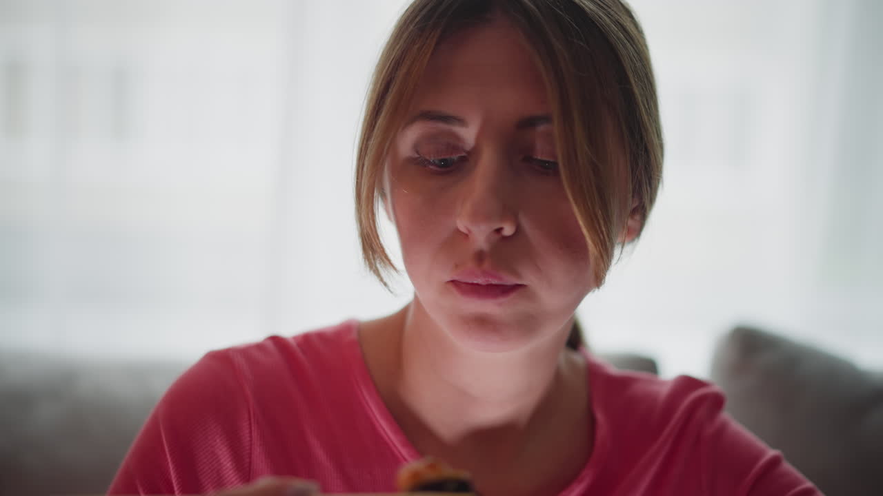 Woman in pink polo shirt seated on couch eating sushi with chopstick in quiet indoor space showing close up of focused face and hand holding sushi with blurred background