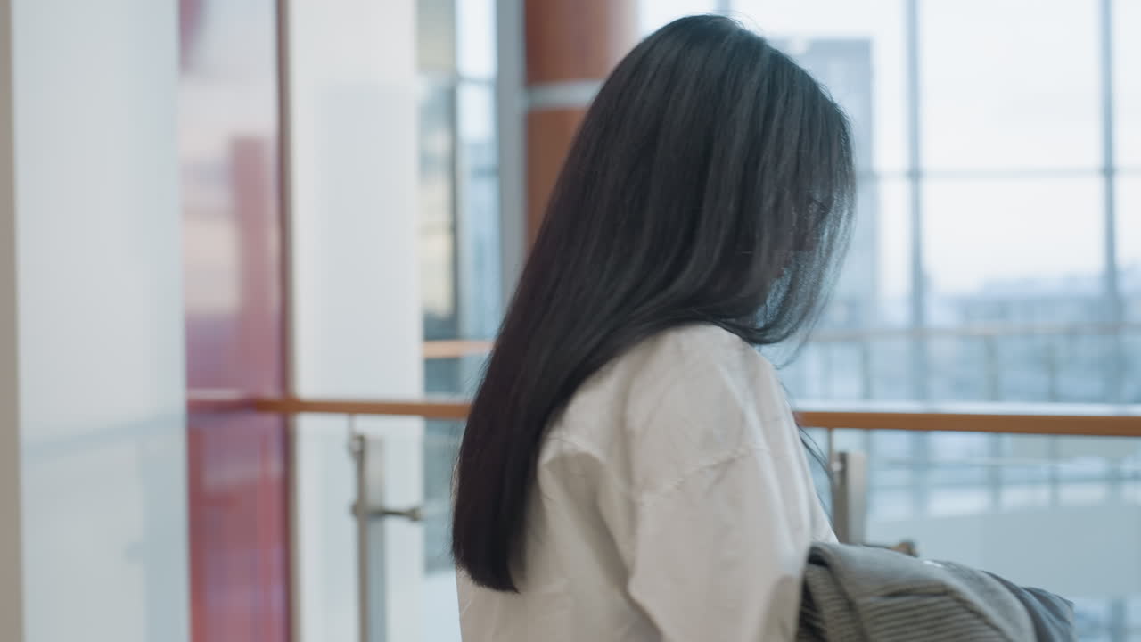 Young lady with long hair holding jacket and book walks indoors near glass railing in sunlit modern mall, preparing to sit as colorful green lights blink in background