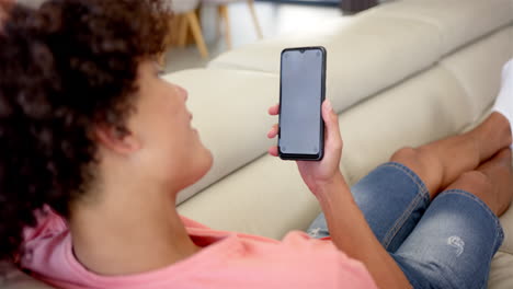 un joven biracial sostiene un teléfono inteligente en un sofá en casa