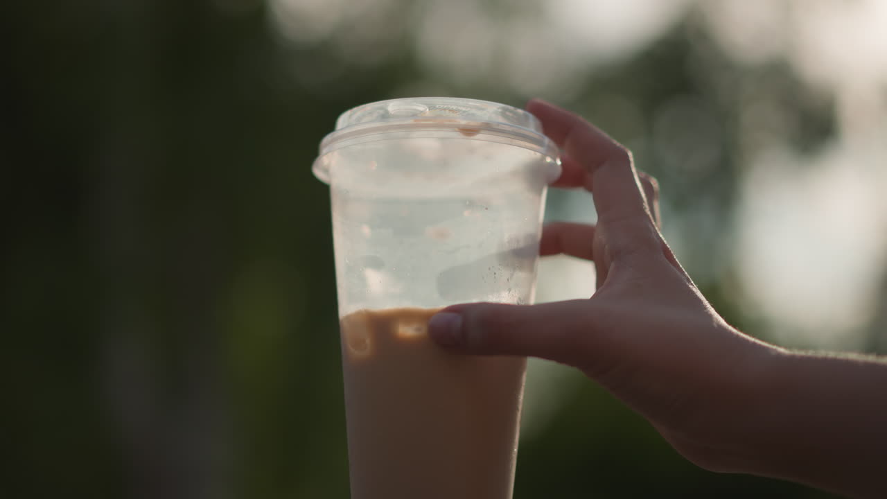 Persona che si gode un caffè freddo con ghiaccio al parco durante il tramonto, mano illuminata dal sole che solleva il caffè freddo in un tranquillo spazio verde urbano, persona rilassata che assapora la bevanda ghiacciata sotto la luce dorata del sole in una vivace area del parco cittadino