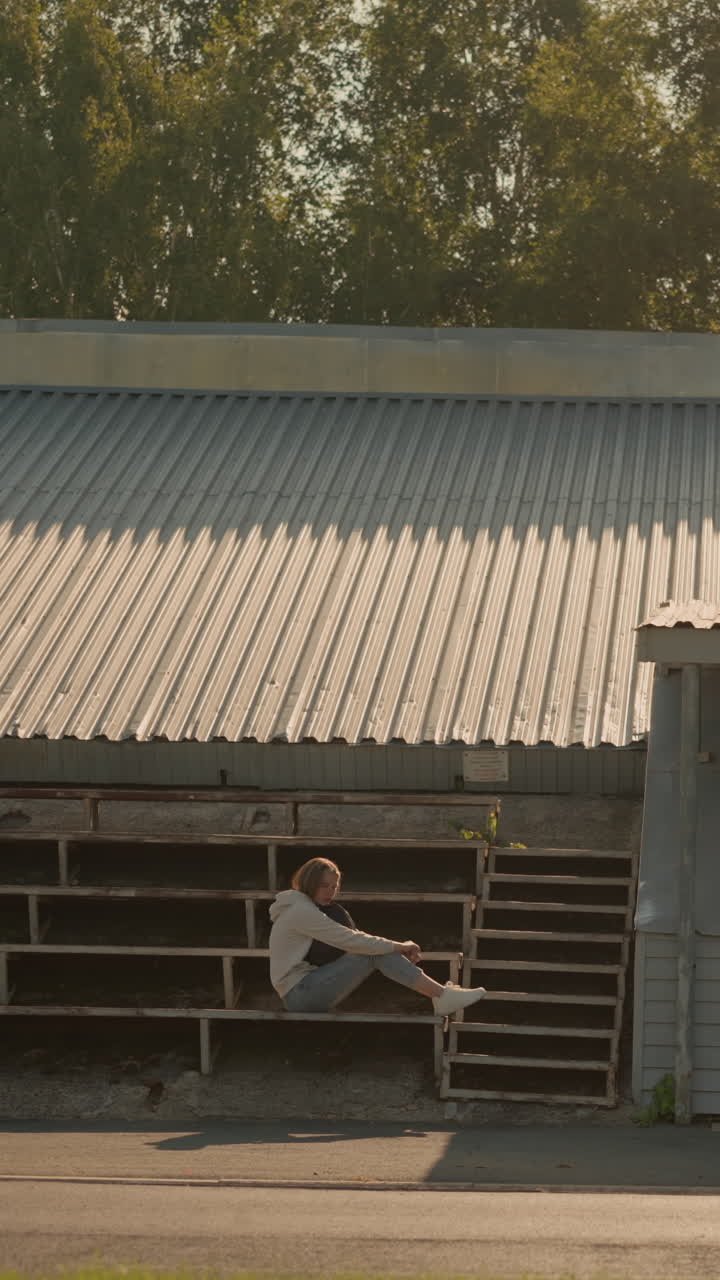 joven sentada sola en las gradas del estadio en una postura pensativa bajo una estructura de techo metálico, rodeada de naturaleza y sombras ligeras de los árboles