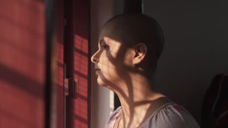 sad and lonely bald indian girl standing near window and looking outside from home due to pandemic covid-19 lockdown