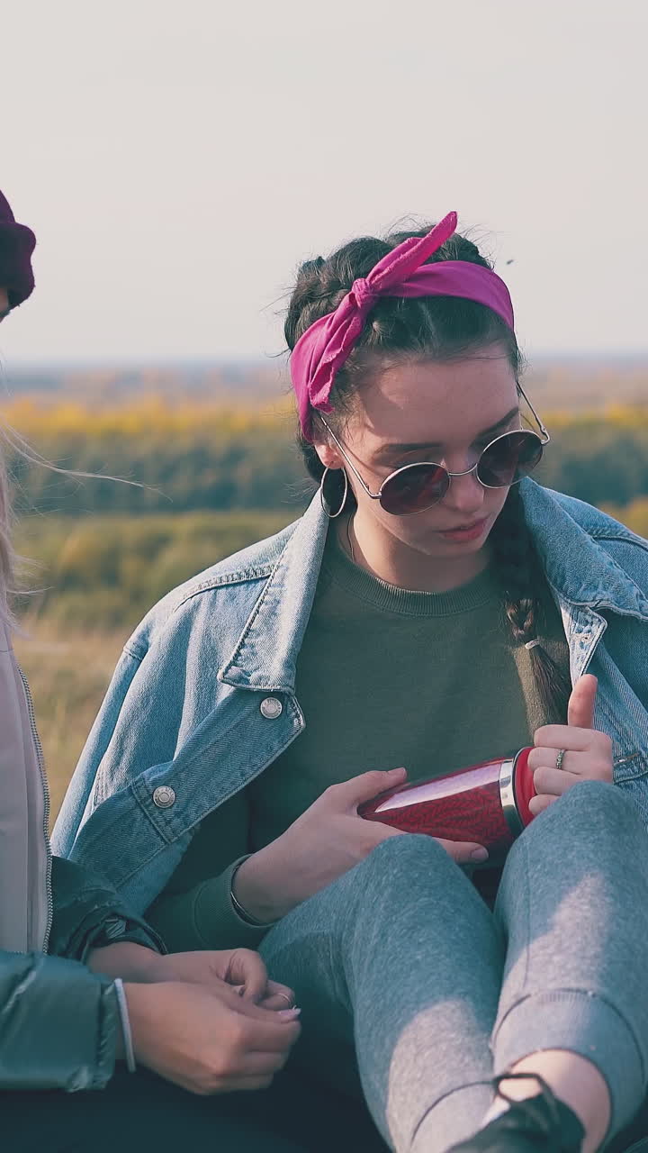 cheerful girls hikers with smartphone and thermos rest on green meadow in warm autumn evening close view