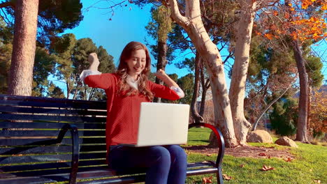 una feliz, atractiva, joven, mujer caucásica en su computadora portátil en el parque, en un banco - lanza sus brazos en el aire para celebrar su logro y ríe
