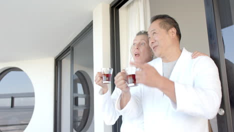 senior couple in bathrobes enjoying drinks on balcony during vacation