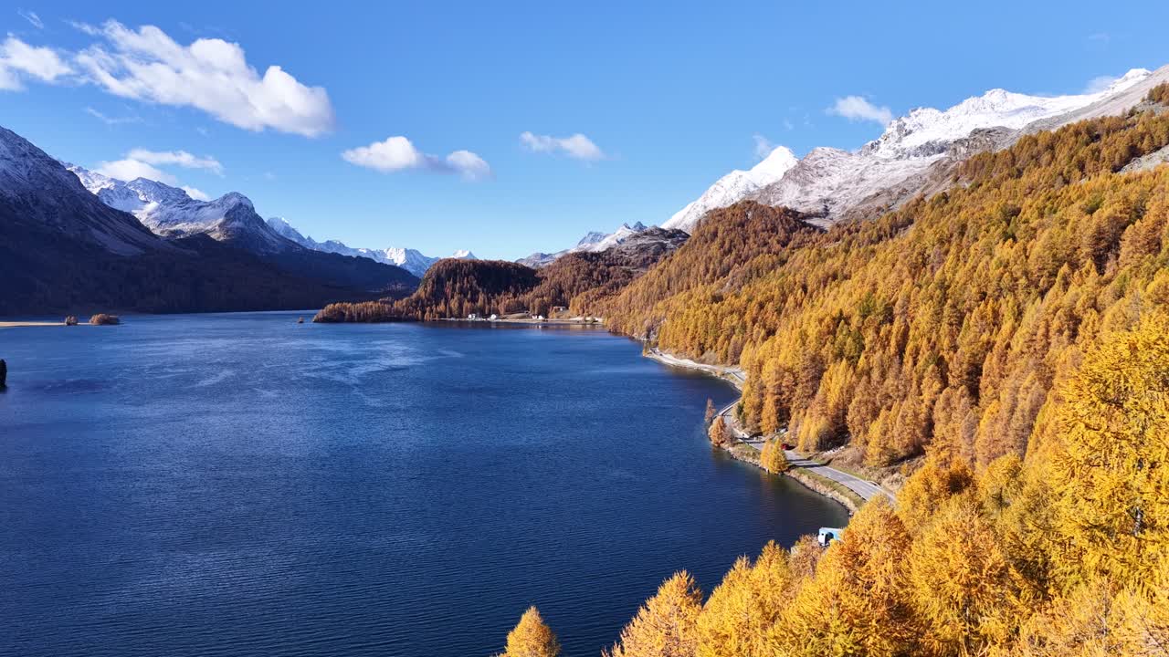Aerial view of a deep blue alpine lake beside golden autumn forests and snowy mountains in the Engadin region
