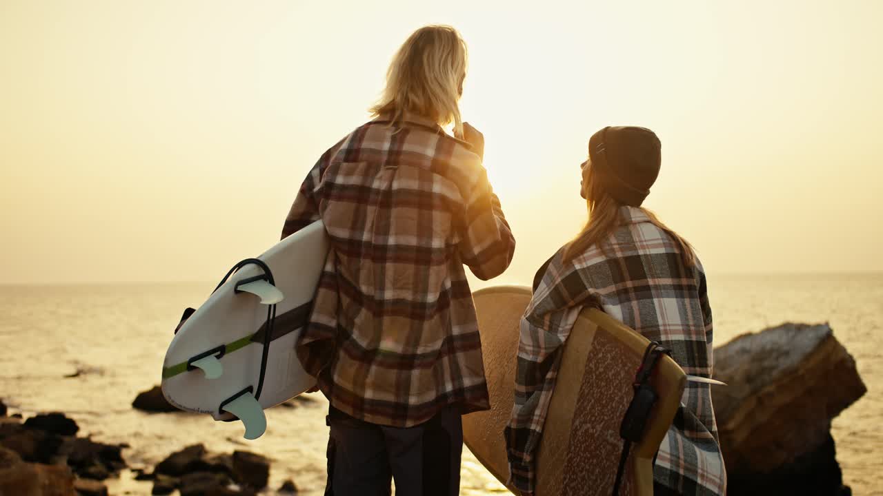 vista trasera de un hombre rubio alto en una camisa a cuadros y su novia rubia en un sombrero y camisa a cuadrados están sosteniendo tablas de surf mientras están de pie en una orilla rocosa cerca del mar y mirando el sol naciente por la mañana en verano