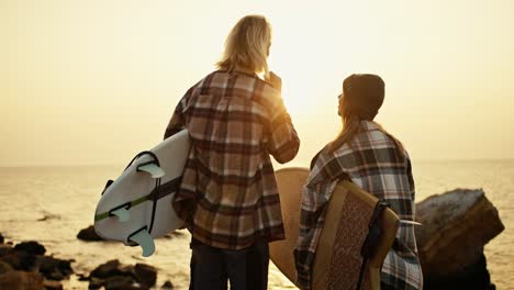 vista trasera de un hombre rubio alto en una camisa a cuadros y su novia rubia en un sombrero y camisa a cuadrados están sosteniendo tablas de surf mientras están de pie en una orilla rocosa cerca del mar y mirando el sol naciente por la mañana en verano