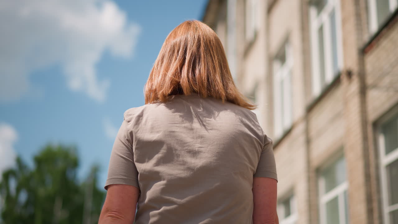 Back view of lady with golden hair walking outdoors under bright blue sky looking right and left while checking time on smartwatch, soft wind moving hair