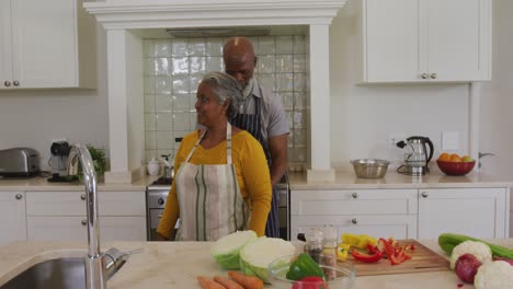 Afroamerikanischer-älterer-Mann,-Der-In-Der-Küche-Zu-Hause-Eine-Schürze-Von-Hinten-An-Seine-Frau-Bindet