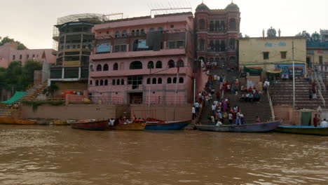 Cinematic-Varanasi-India-Ganges-River-cruise-canal-boat-Northern-State-large-gathering-at-shore-Ancient-Holy-city-Ghat-Pradesh-Province-landscape-gray-cloudy-Holy-muddy-brown-afternoon-sunset-follow