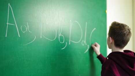 schoolboy writing on blackboard. teen boy writes alphabet on green school chalkboard. backside view of a student with a chalk in the classroom. education concept.