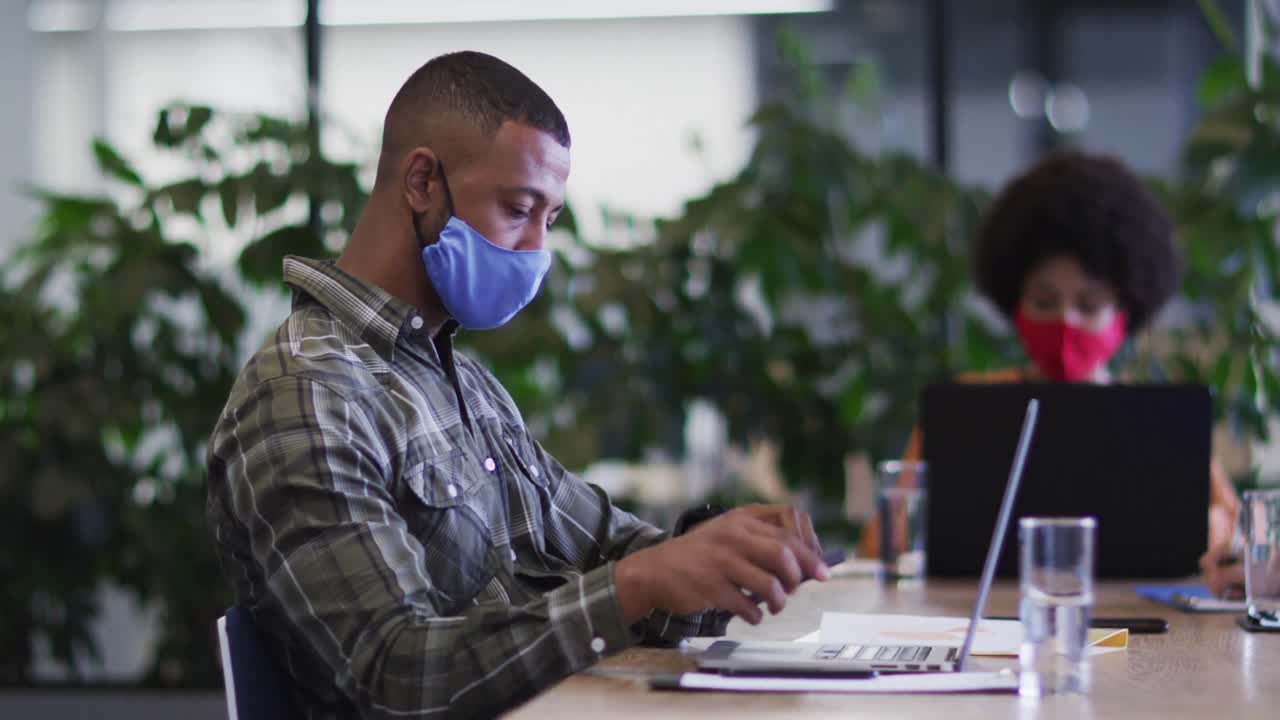 Diverse business people wearing face masks sitting using laptops going through paperwork in office
