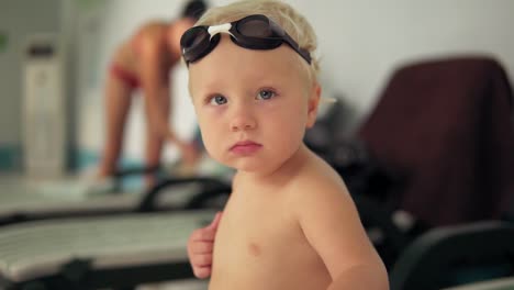 retrato de un hermoso niño pequeño con gafas sentado junto a la piscina y mirando a la cámara esperando a que venga su madre