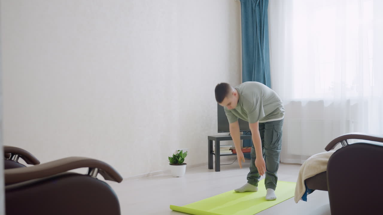 Focused young boy bends forward during stretching routine on green exercise mat in sunlit living room. Casual outfit, soft light, and indoor setting