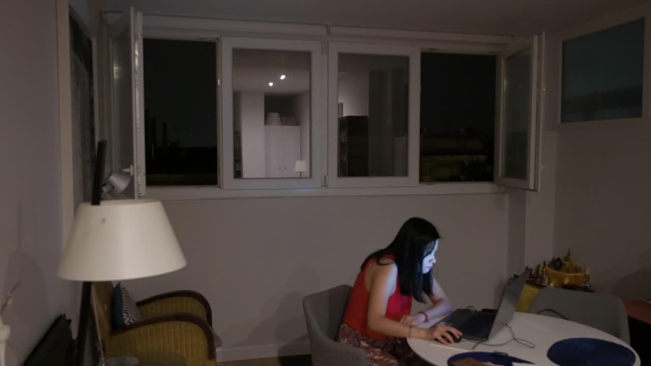 Una mujer usando un portátil en interiores mientras una tormenta eléctrica ocurre afuera, con destellos visibles a través de las ventanas de la casa.