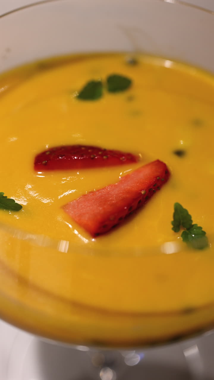 Primo piano di budino di chia al mango cremoso servito in un elegante bicchiere con fette di fragola ed erbe fresche. Verticale.