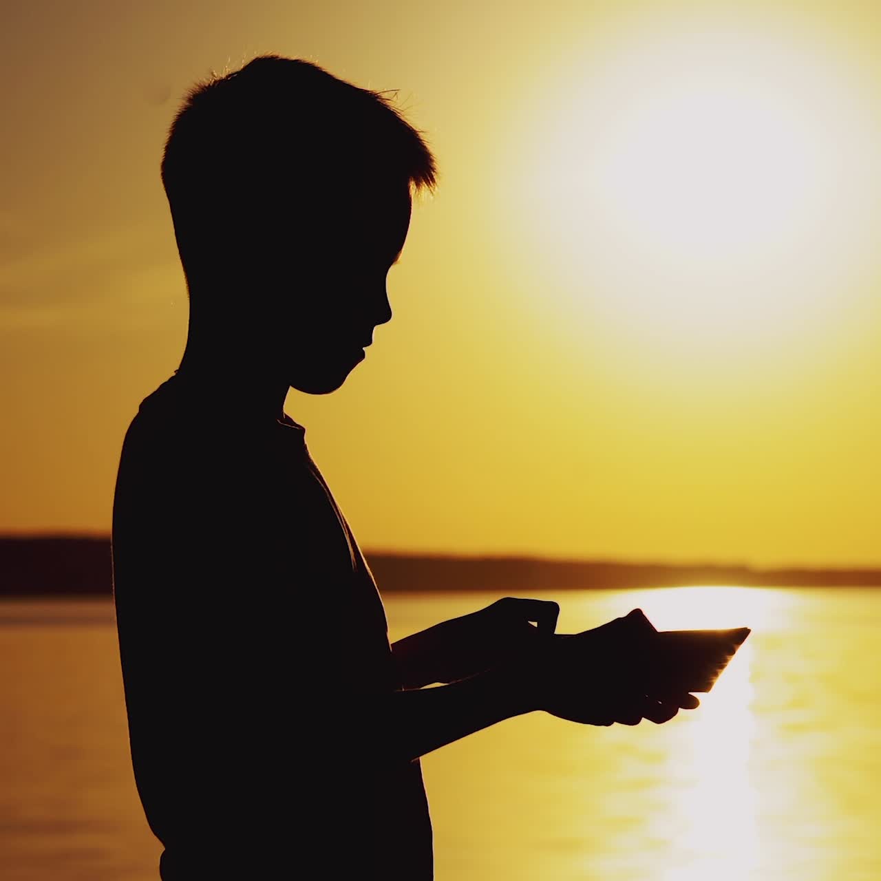 boy is making origami in the form of a ship on the background of sunset by the river in the summer