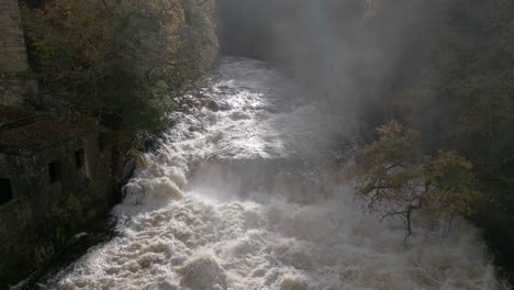 slow motion drone footage descends close to a fast flowing river and waterfall surrounded by a forest of broadleaf trees in autumn