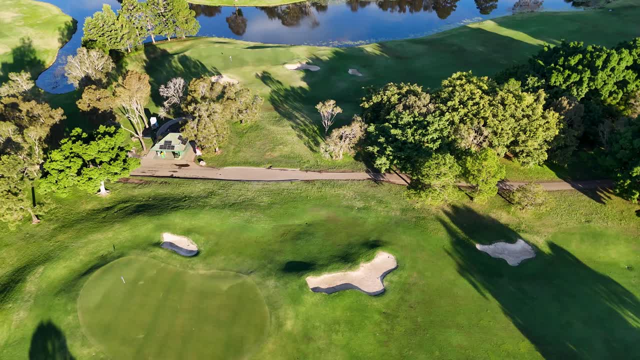 Drone footage captures a lush golf course with vibrant greens, sand bunkers, and a serene water hazard under bright sunlight