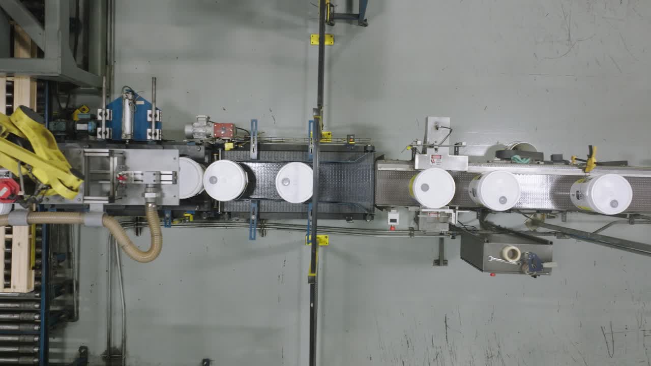 Top-down aerial shot of a yellow robotic arm performing palletizing by stacking white pails or buckets onto wooden pallets on an automated conveyor belt in a manufacturing factory