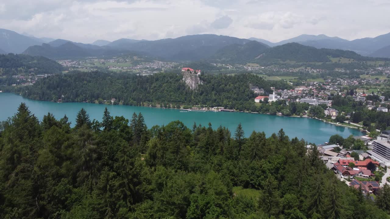 Ripresa aerea rivelatrice di una foresta verde con il Lago di Bled naturale e la catena montuosa sullo sfondo durante una giornata nuvolosa, Slovenia.
