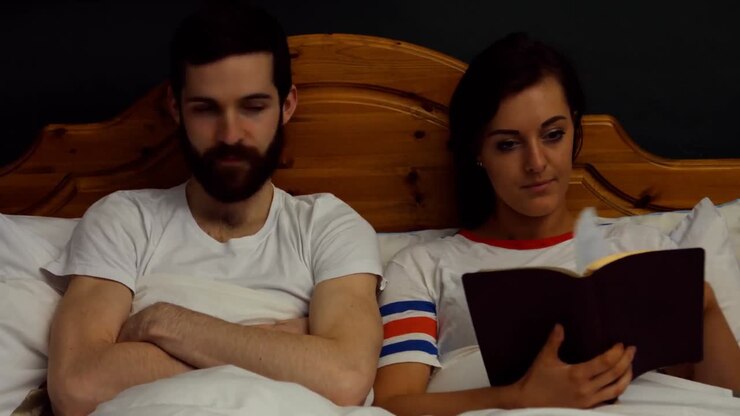 Man getting annoyed while woman reading book on bed