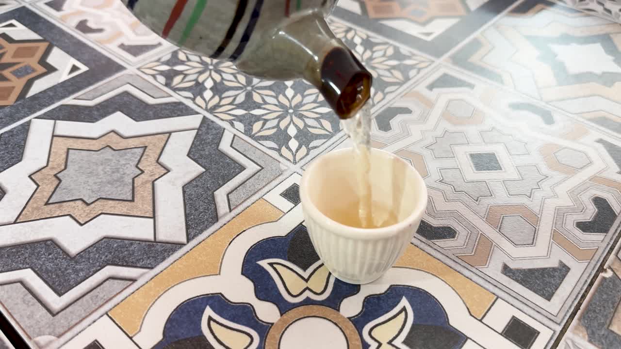 Hand pours hot tea from striped teapot into cup on colorful tile table, natural daylight