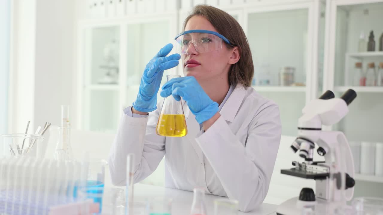 A female scientist in safety glasses and gloves examining a liquid in a flask in a laboratory