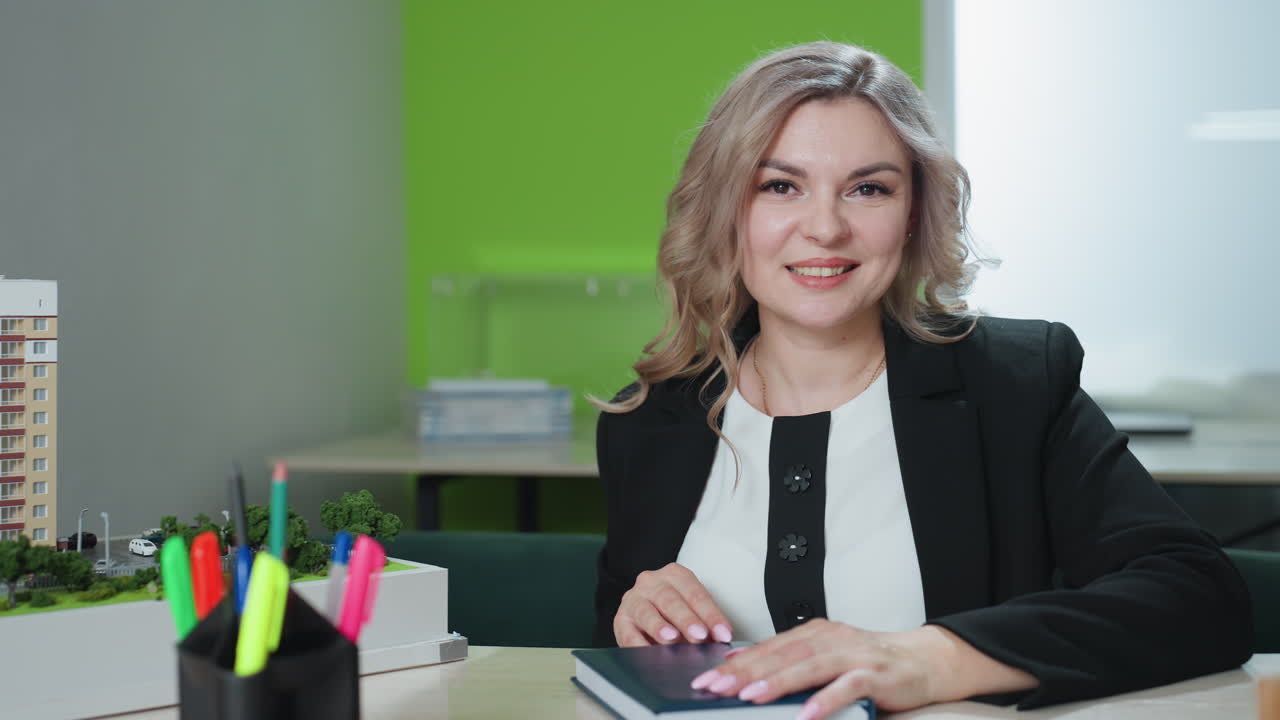 real estate agent closes book smiling at desk with architectural model laptop plant colorful pens in bright office setting professional poised organized workspace details showcase confidence