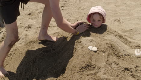 ein mädchen genießt es, am strand im sand begraben zu werden, während jemand ihrer sandschöpfung den letzten schliff hinzufügt