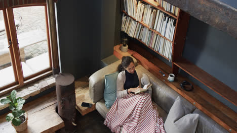 biracial woman lying on sofa under blanket and reading book at home, slow motion