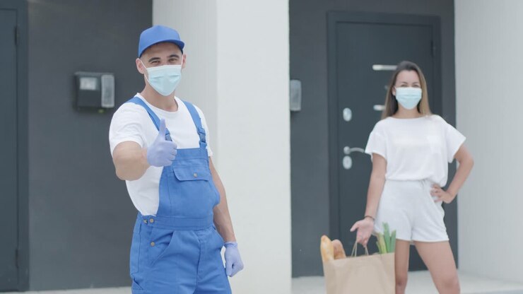 Beautiful woman with protective mask meets a delivery man wearing mask and gloves who gives her a shopping package with food beside her home with safety. Food delivery, home delivery in quarantine