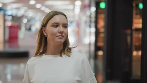 compradora femenina en una camiseta blanca caminando por un centro comercial, mirando algo en el fondo, rodeada de luces de colores y un fondo borroso con objetos poco claros