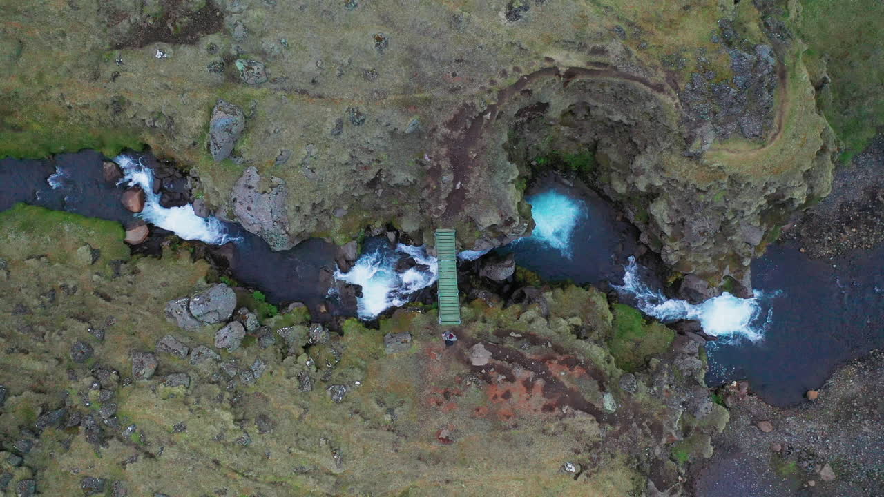 vista superior de un hombre cruzando un pequeño puente de madera a través de un arroyo que fluye en el sur de islandia - drone aéreo