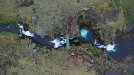 Vista-Superior-De-Un-Hombre-Cruzando-Un-Pequeño-Puente-De-Madera-A-Través-De-Un-Arroyo-Que-Fluye-En-El-Sur-De-Islandia---Drone-Aéreo