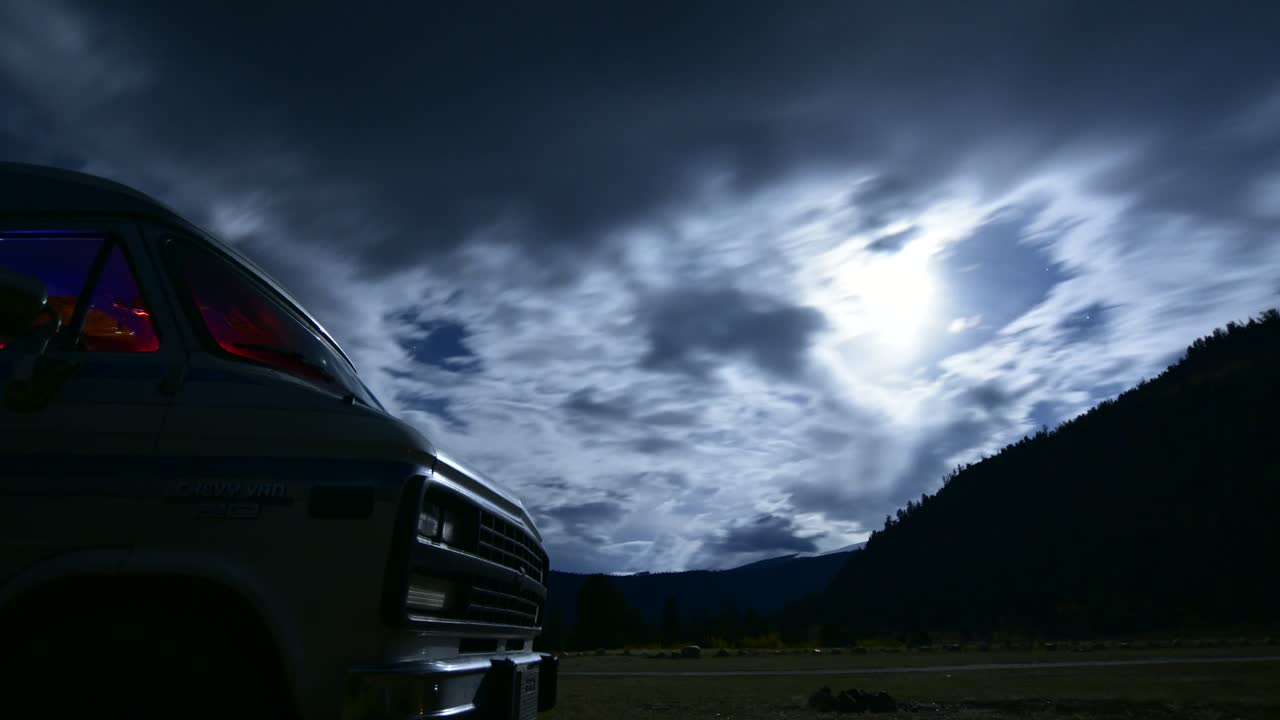 Campers flash colorful lights in their van while dramatic clouds drift by the bright moon in this eerie time lapse scene - Chevy Van