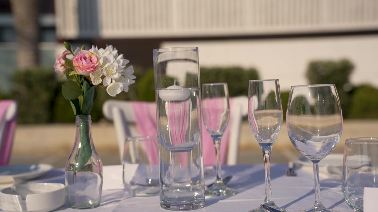 A table is arranged with glasses and flowers for an outdoor meal at a sunny location. The setup includes a candle in a tall glass. People will sit down soon.
