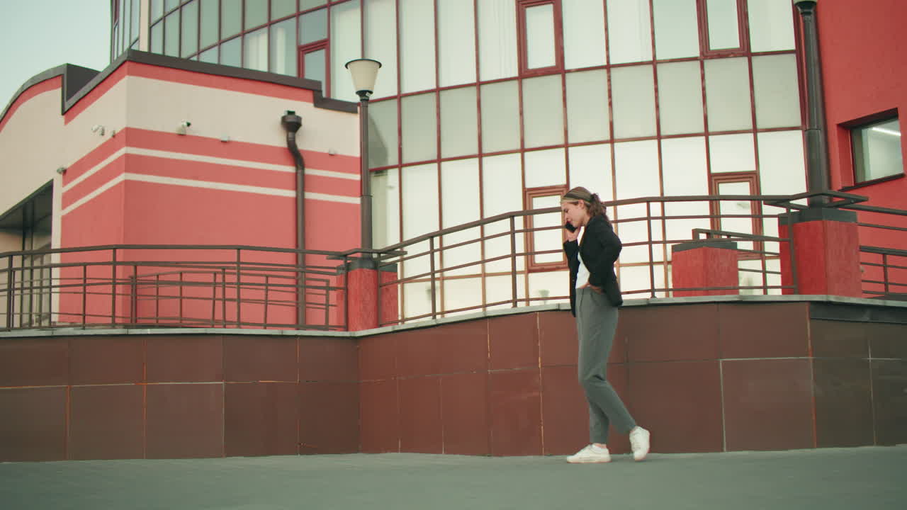 Woman on phone call walking near fence in modern urban environment, dressed in professional attire, appearing focused and engaged, with building reflections in glass windows