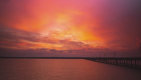 Epic-cloudscape-of-brilliant-orange-red-hues-at-sunset-over-river-by-highway-bridge