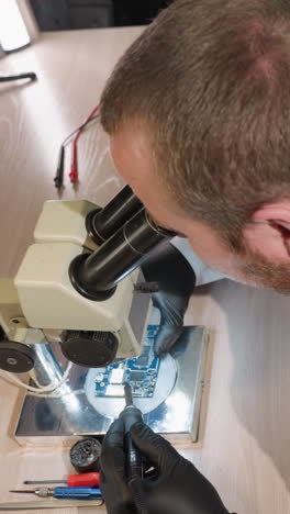 una vista hacia abajo de un técnico en una bata de laboratorio blanca trabajando en una placa de circuito bajo un microscopio, el espacio de trabajo está equipado con varias herramientas