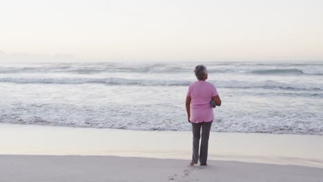 Eine-Afroamerikanische-Seniorin-Hält-Eine-Yogamatte-Und-Blickt-Auf-Die-Wellen,-Während-Sie-Am-Strand-Steht