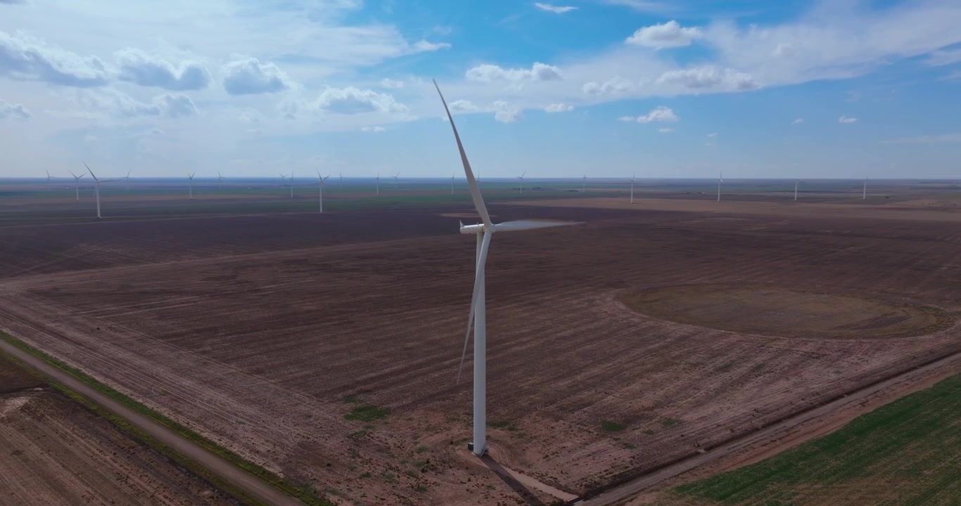 Un drone si muove lentamente attorno a una singola turbina eolica isolata, mantenendola centrata sopra una pianura agricola secca del Texas, con decine di turbine visibili all'orizzonte